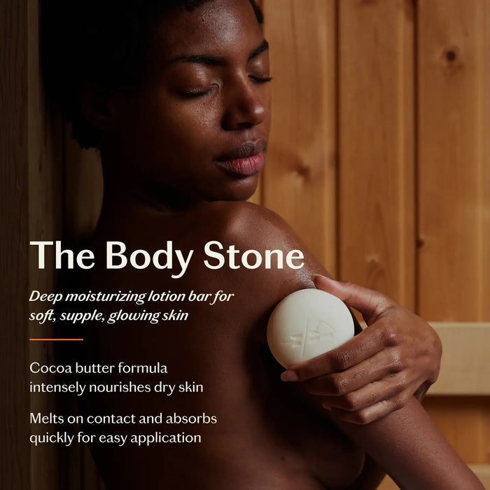 A round white KATE MCLEOD Grounding Stone deep moisturizing lotion bar sits on textured fabric next to an open container with a wooden lid, featuring a dark green label with orange and white text, highlighting its luxurious blend of cocoa butter concentrate and natural essential oils.