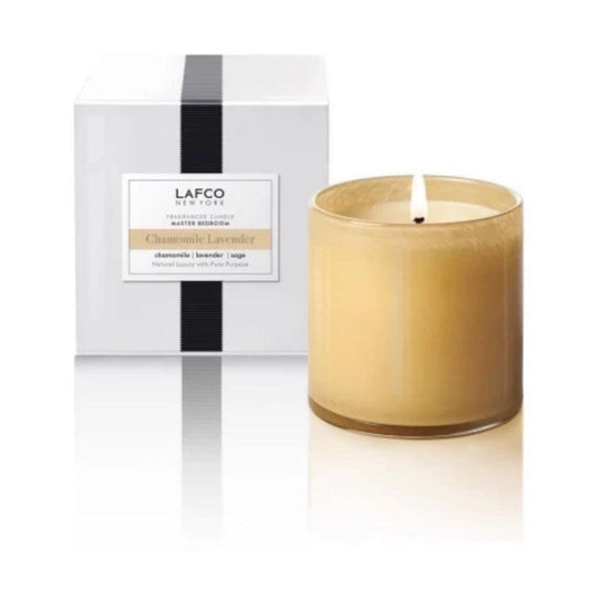 A lit beige luxury candle in a glass holder sits beside a white LAFCO CHAMOMILE LAVENDER, BEDROOM candle box featuring a black vertical stripe and branded label.