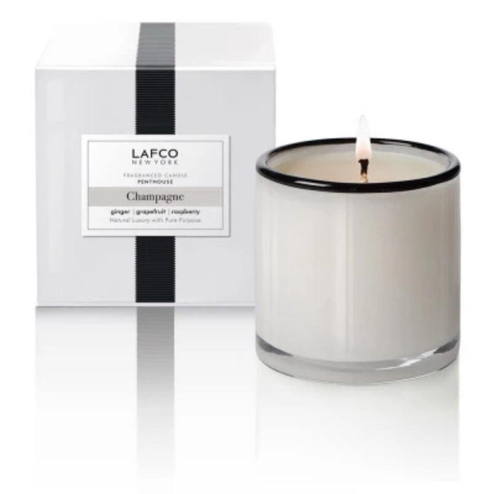 A white LAFCO art glass candle with a black rim, "CHAMPAGNE, PENTHOUSE 15OZ," burns next to its minimalist white box featuring a black vertical stripe and the scent notes: ginger, grapefruit, and raspberry.