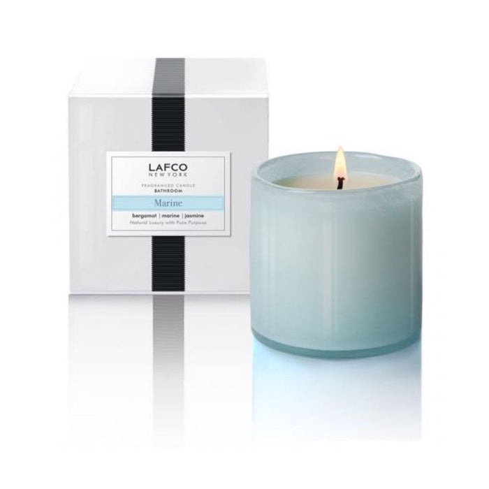 A LAFCO - MARINE, BATHROOM 15OZ candle glows in a light blue art glass vessel, with its white box featuring a black stripe and matching label visible in the background.