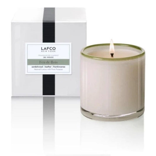 A lit LAFCO Ski House Feu de Bois candle in art glass sits next to a white LAFCO New York box. Enjoy notes of sandalwood, leather, and frankincense—a classic luxury scent.