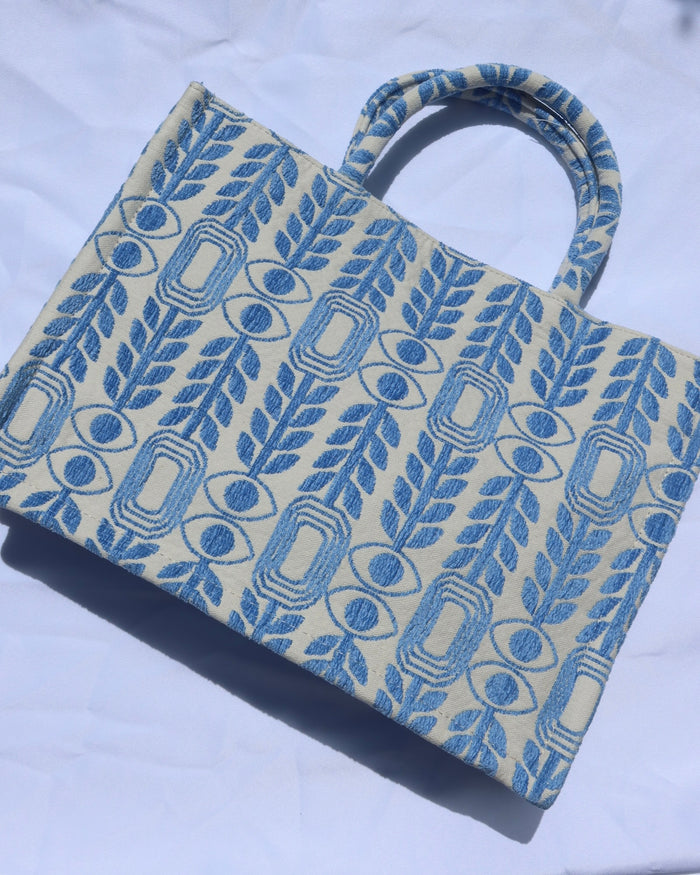 large handbag with woven light blue motif featuring evil eyes, olive branches, and abstract shapes handcrafted in Greece. Greek artisinal handbag as seen on Emily in Paris