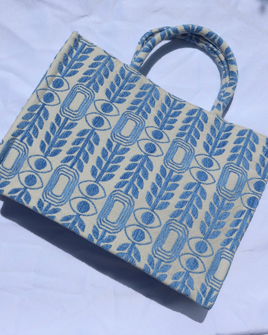 large handbag with woven light blue motif featuring evil eyes, olive branches, and abstract shapes handcrafted in Greece. Greek artisinal handbag as seen on Emily in Paris