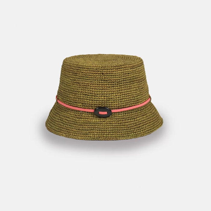 The SANS ARCIDET LEO BUCKET HAT is a handmade pink raffia sun hat for women from Madagascar, featuring a blue band and black oval accent, shown against a plain light gray background.