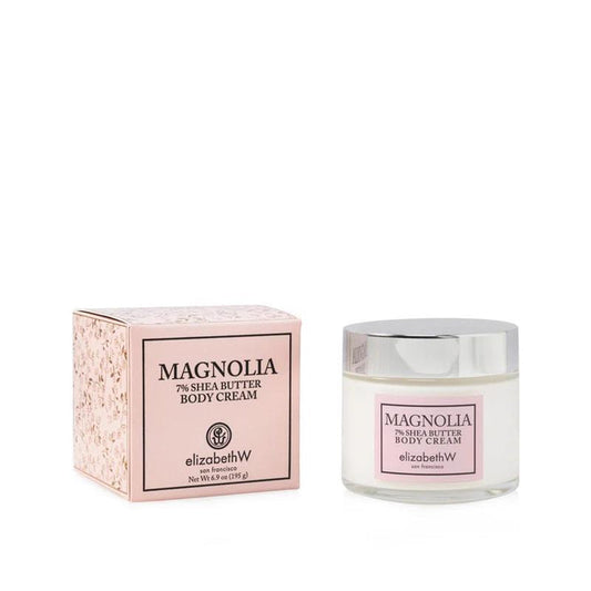 A jar of E WIGHTMAN & CO MAGNOLIA BODY CREAM with a silver lid sits beside its matching floral light pink box—an intensely hydrating botanical cream.
