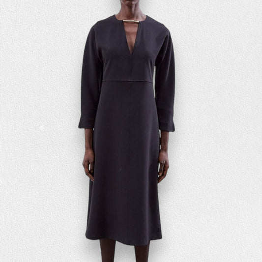 A model wears the MEIMEIJ Long Sleeve Dress with Metallic Collar—a black, midi-length dress with a V-neckline by MEIMEIJ—standing against a textured light background.