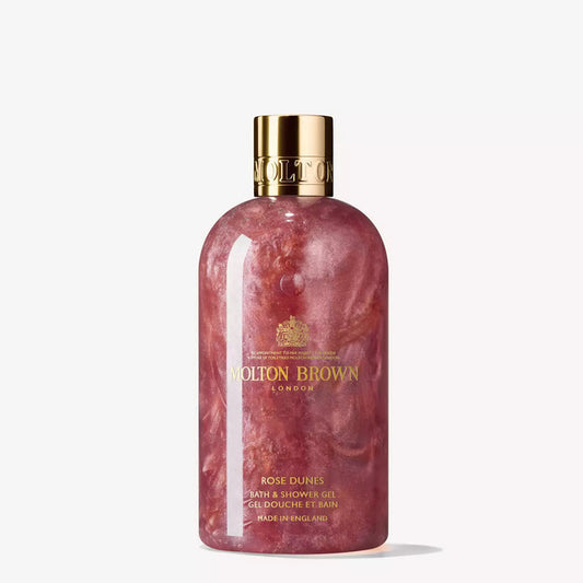 A bottle of MOLTON BROWN ROSE DUNES SHOWER GEL with ambery, shimmering pink liquid and a gold cap is displayed against a white background.