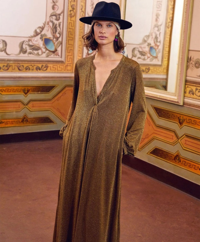 A woman wearing the MOMONI - AMEDE DRESS IN LUREX JERSEY and a black wide-brimmed hat stands in an ornately decorated room with large mirrors and intricate wall designs.
