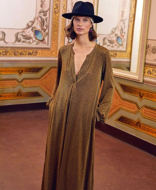A woman wearing the MOMONI - AMEDE DRESS IN LUREX JERSEY and a black wide-brimmed hat stands in an ornately decorated room with large mirrors and intricate wall designs.