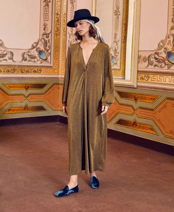 A woman wearing the MOMONI - AMEDE DRESS IN LUREX JERSEY and a black wide-brimmed hat stands in an ornately decorated room with large mirrors and intricate wall designs.