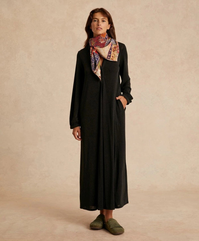 A woman wearing the MOMONI - AMEDE DRESS IN LUREX JERSEY and a black wide-brimmed hat stands in an ornately decorated room with large mirrors and intricate wall designs.