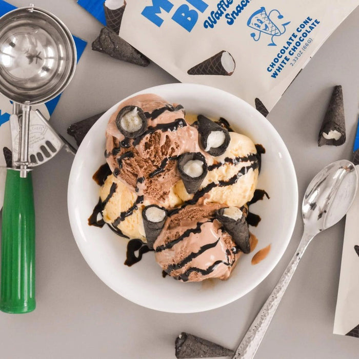 A person dressed in a light blue outfit sits on a plush, royal blue rug holding a bag of Muddy Bites Cookies & Cream Waffle Cones in one hand and a small white chocolate waffle cone snack in the other. The beige and blue bag features an illustration of chocolate-dipped waffle cones.
