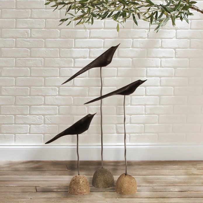 The NAPA HOME AND GARDEN "THE FLOCK" Set of 3-Black minimalist bird sculptures are displayed indoors on slender stands with stone bases, set on a wooden floor against a white brick wall with leafy branches above.