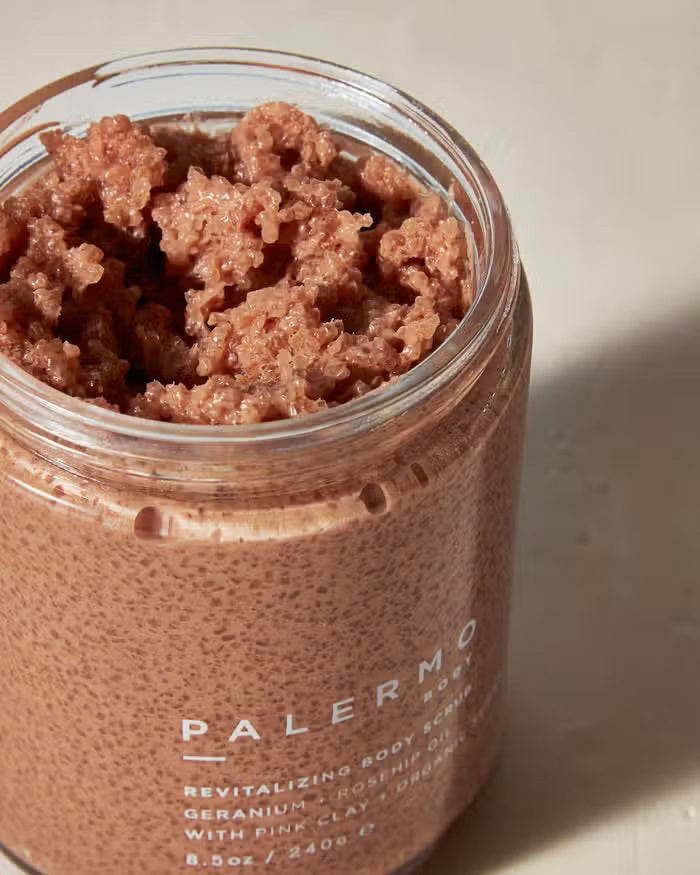 A clear jar with a black lid holds Palermo Body's Geranium and Rose Hip Body Scrub, made with geranium, rosehip oil, French pink clay, and organic sugar. Labeled 8.5 oz (240g).
