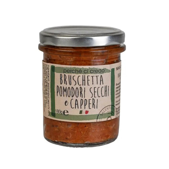 A jar of Zia Pia Imports' PERCHE CI CREDO - SUN-DRIED TOMATO & CAPER BRUSCHETTA, featuring sun-dried tomatoes, capers, and extra virgin olive oil. The label is beige with green and red accents, displays text in Italian, and indicates the product weight of 180 grams. The jar comes with a metal lid and is perfect for lovers of authentic Italian foods.