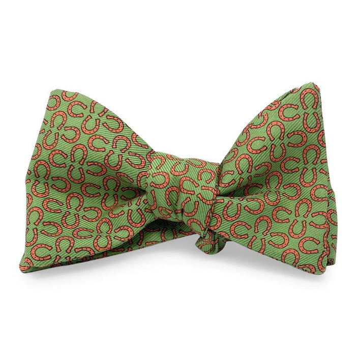 The BIRD DOG BAY PETER-BLAIR RINGERS Silk Bow Tie in green features a handmade design with small red horseshoes, an adjustable neck, and is displayed neatly tied against a white background.
