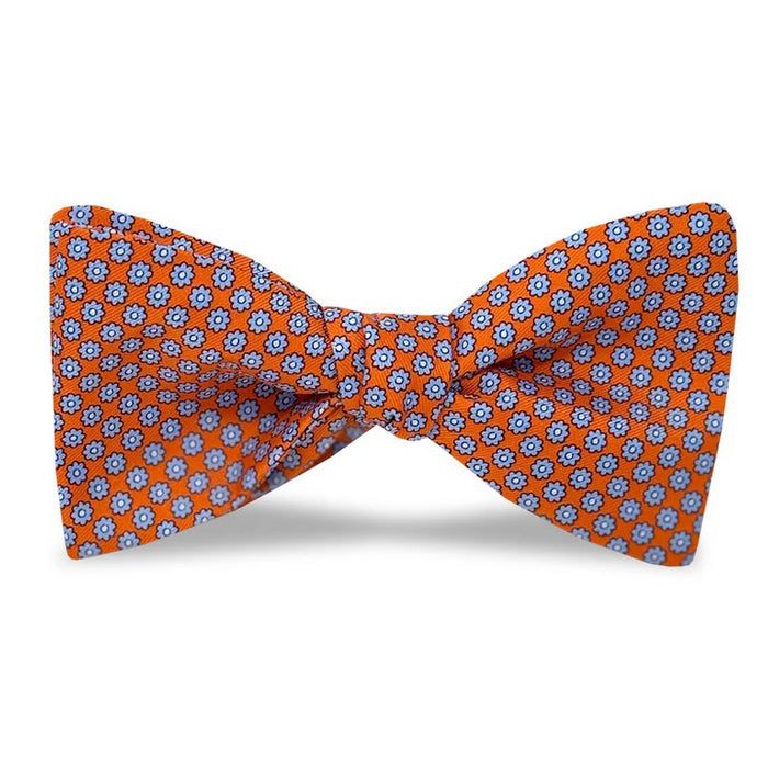 The BIRD DOG BAY PETER-BLAIR - WYTHE SILK BOW TIE IN CORAL features a repeating blue and white floral pattern, is handmade in North Carolina with an adjustable neck, and is shown against a plain white background.