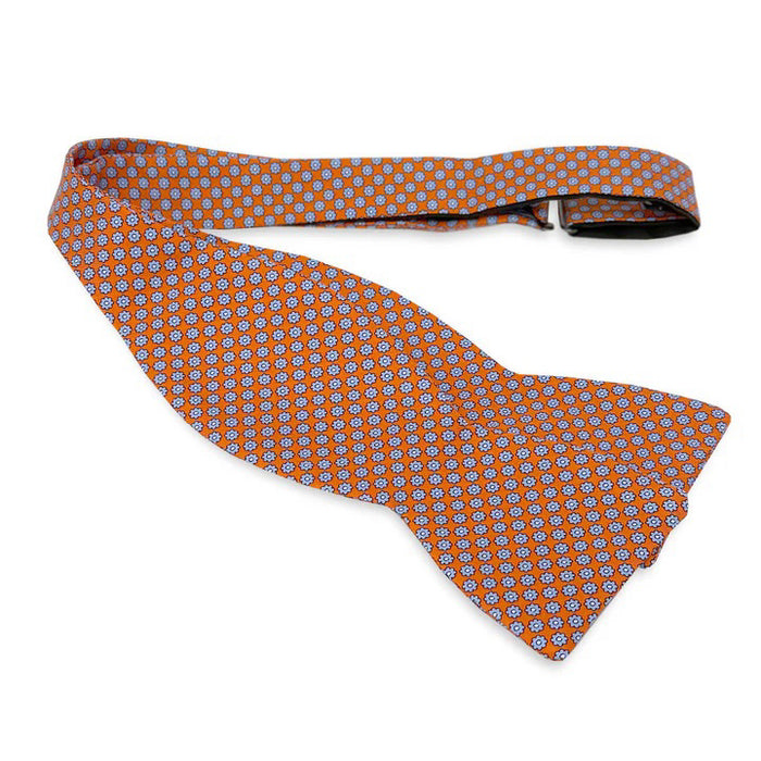 The BIRD DOG BAY PETER-BLAIR - WYTHE SILK BOW TIE IN CORAL features a repeating blue and white floral pattern, is handmade in North Carolina with an adjustable neck, and is shown against a plain white background.