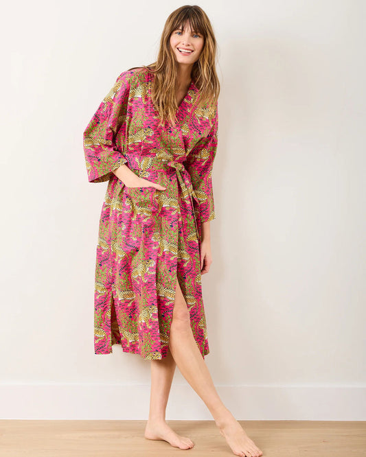 A smiling person with long hair wears the PRINTFRESH - BAGHEERA COTTON ROBE IN HOT PINK, hand screen-printed by PRINTFRESH, standing barefoot on a wooden floor against a plain light-colored wall.
