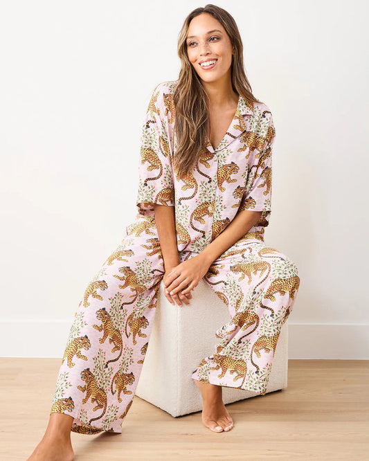 A woman sits barefoot on a white cube against a plain white wall and light wood floor, smiling as she wears the PRINTFRESH - BAGHEERA SATIN WILDEST DREAM PAJAMA SET IN BLUSH by PRINTFRESH—a light pink leopard print pajama set crafted from eco-friendly viscose.