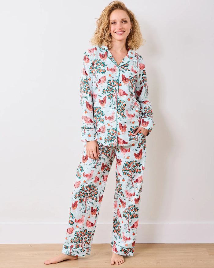 A person stands barefoot against a white wall in PRINTFRESH's pink organic cotton HEN HOUSE PAJAMA SET, featuring hand screen-printed roosters and floral patterns. The pajamas have long sleeves and pants, with one hand in a pocket and their head slightly tilted.