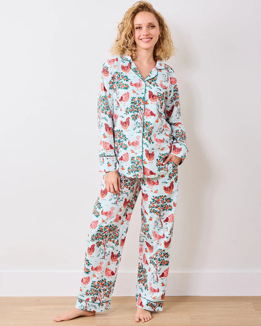 A person stands barefoot against a white wall in PRINTFRESH's pink organic cotton HEN HOUSE PAJAMA SET, featuring hand screen-printed roosters and floral patterns. The pajamas have long sleeves and pants, with one hand in a pocket and their head slightly tilted.