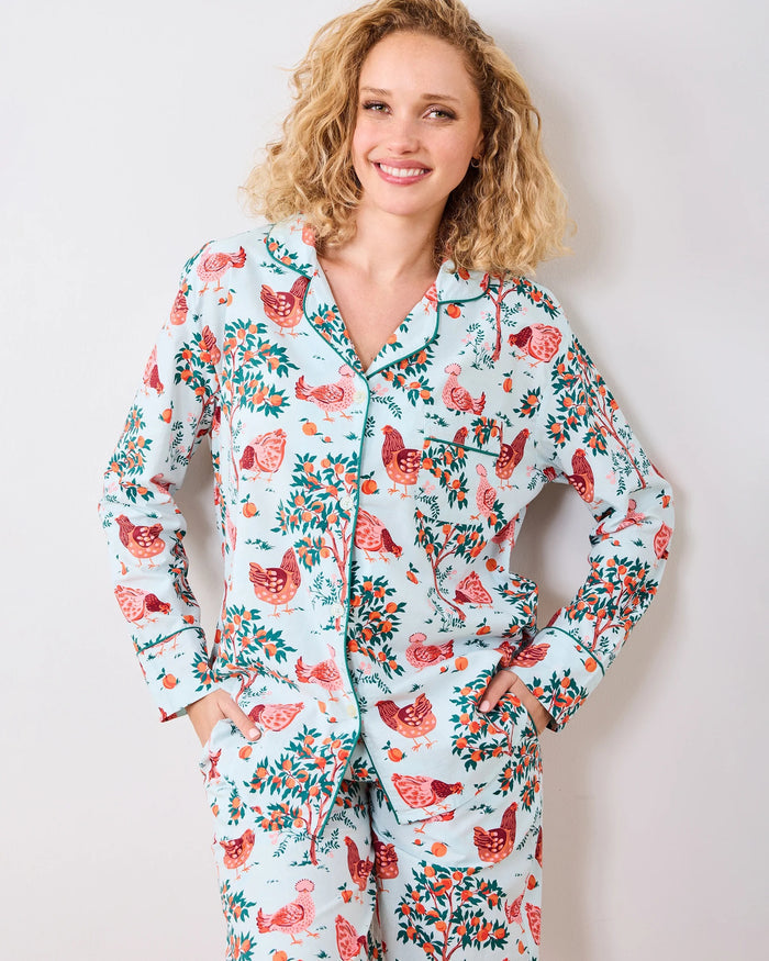 A person stands barefoot against a white wall in PRINTFRESH's pink organic cotton HEN HOUSE PAJAMA SET, featuring hand screen-printed roosters and floral patterns. The pajamas have long sleeves and pants, with one hand in a pocket and their head slightly tilted.