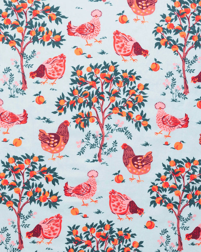A person stands barefoot against a white wall in PRINTFRESH's pink organic cotton HEN HOUSE PAJAMA SET, featuring hand screen-printed roosters and floral patterns. The pajamas have long sleeves and pants, with one hand in a pocket and their head slightly tilted.