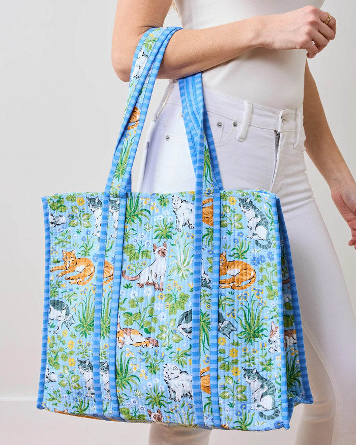 A person in white clothes holds the PRINTFRESH - MUST LOVE CATS TRAVELER TOTE, a light blue quilted cotton bag featuring cats among flowers and greenery. With its spacious design and long handles, it's ideal for any cat lover.