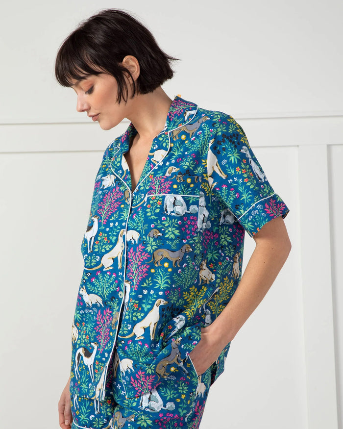 A person stands against a plain white wall wearing the stylish PRINTFRESH - MUST LOVE DOGS PAJAMA SHORT SET made of organic cotton. The set, adorned with colorful dog and floral illustrations on a blue background, features pockets where the person's hands rest.