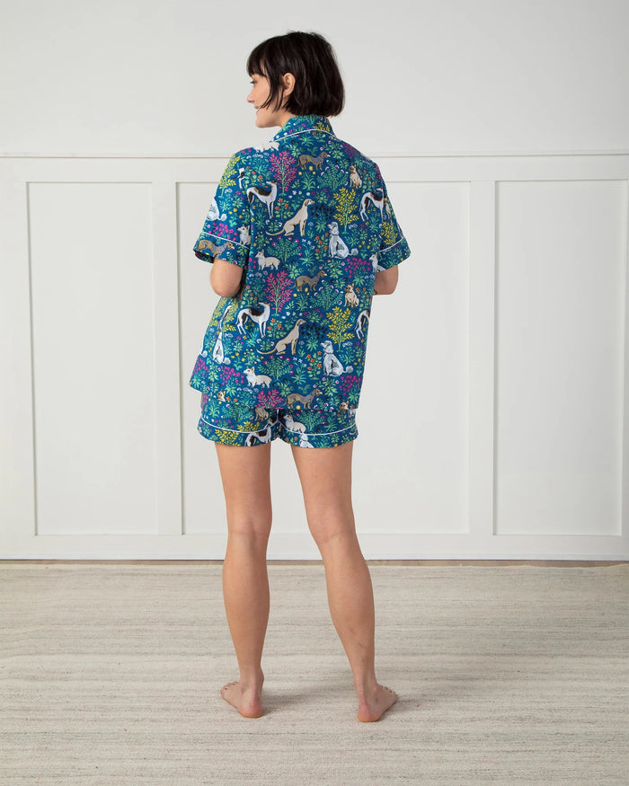 A person stands against a plain white wall wearing the stylish PRINTFRESH - MUST LOVE DOGS PAJAMA SHORT SET made of organic cotton. The set, adorned with colorful dog and floral illustrations on a blue background, features pockets where the person's hands rest.