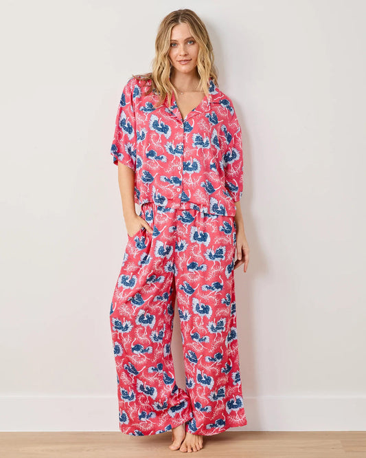 A woman with wavy blonde hair stands barefoot against a white wall, smiling softly as she models the PRINTFRESH - OSTRICH DANCE SATIN WILDEST DREAM PAJAMA SET, a matching pink pajama set by PRINTFRESH featuring a blue and white floral pattern. This eco-friendly satin sleepwear combines stylish design and sustainable comfort for your wildest dreams.
