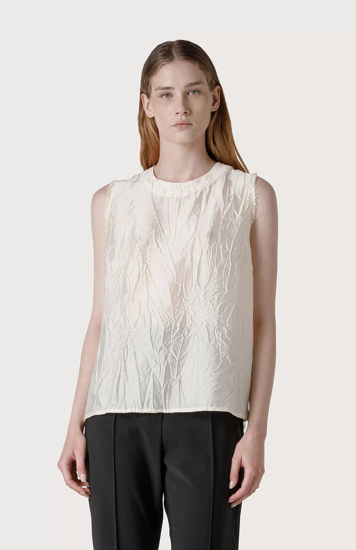 A woman with long, straight brown hair wears a SEVENTY SLEEVELESS TOP in textured off-white creased fabric and black pants, standing against a plain light gray background.