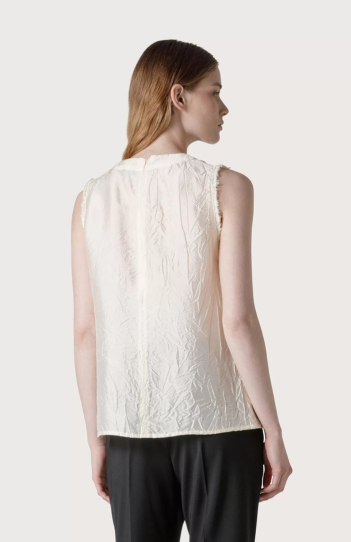 A woman with long, straight brown hair wears a SEVENTY SLEEVELESS TOP in textured off-white creased fabric and black pants, standing against a plain light gray background.