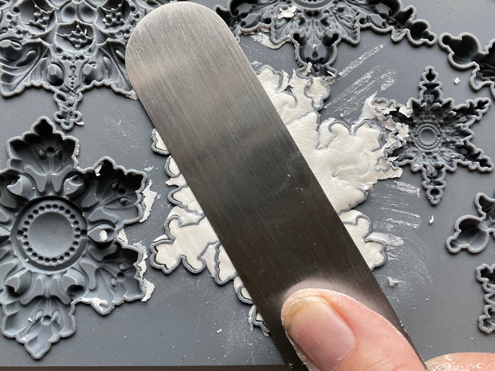 A close-up of a hand using a flat metal spatula to spread white adhesive or putty over a dark gray surface decorated with ornate snowflake and floral embossed designs.