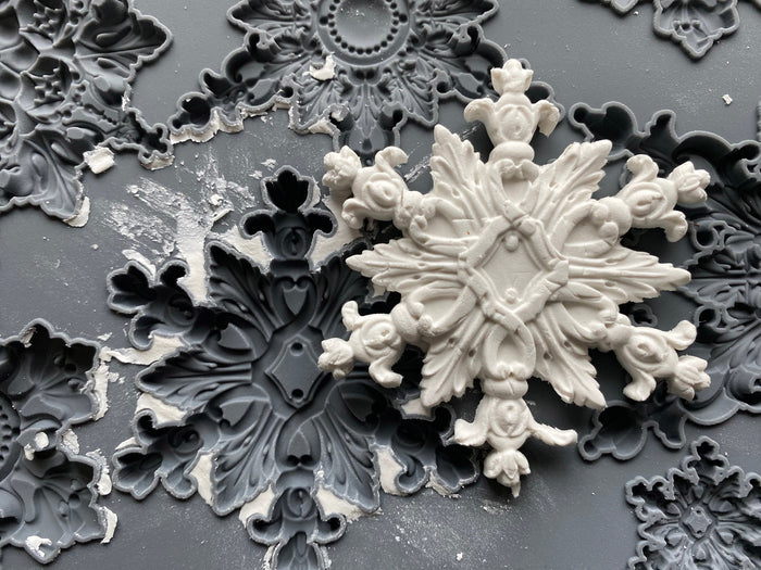 A close-up of a white ornate snowflake-shaped decorative object resting on a dark gray surface with matching gray ornamental molds or trays.