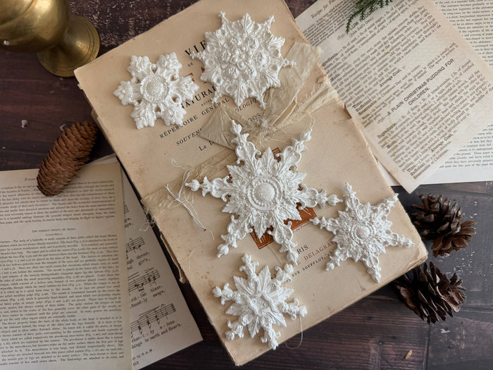 Five white crocheted snowflake ornaments arranged on an old book, surrounded by vintage pages, pinecones, and a brass object on a dark wooden surface.