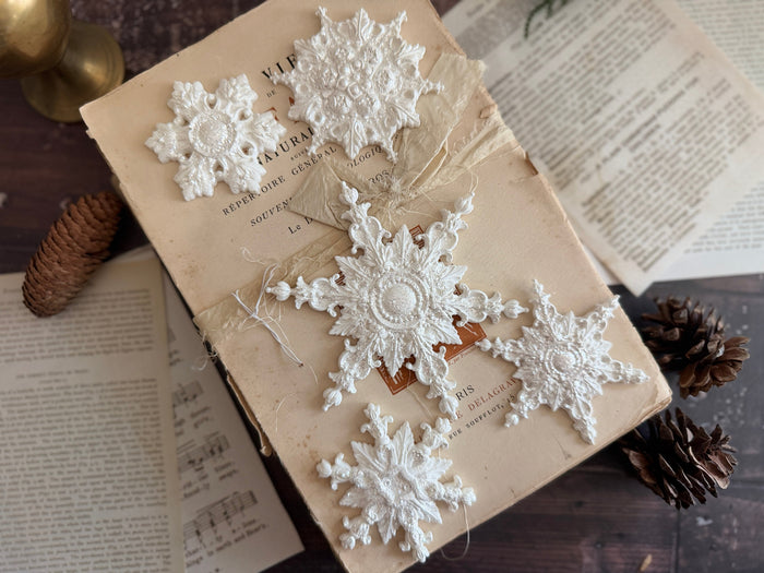 Five white crocheted snowflake ornaments arranged on an antique beige book, surrounded by open vintage pages, pinecones, and a brass candle holder on a dark wooden surface.