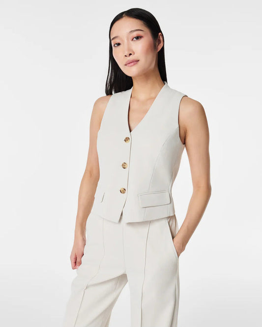 A person with long dark hair models the SPANX - CAREFREE CREPE VEST, a sleeveless, light beige tailored vest with gold buttons, paired with matching high-waisted trousers, standing hands in pockets against a plain white background.