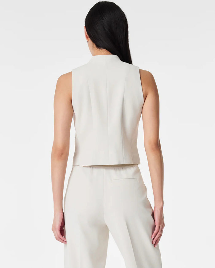 A person with long dark hair models the SPANX - CAREFREE CREPE VEST, a sleeveless, light beige tailored vest with gold buttons, paired with matching high-waisted trousers, standing hands in pockets against a plain white background.