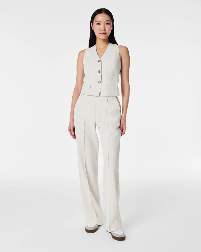 A person with long dark hair models the SPANX - CAREFREE CREPE VEST, a sleeveless, light beige tailored vest with gold buttons, paired with matching high-waisted trousers, standing hands in pockets against a plain white background.