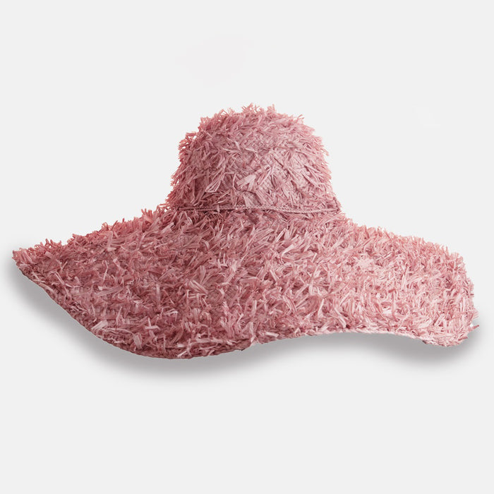 The SUMMERTIME HAT by SANS ARCIDET is a wide-brimmed, floppy raffia hat from Madagascar, featuring a rough, shaggy texture and uneven edges, shown against a plain white background.