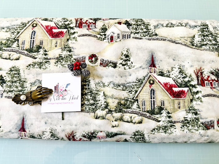 A close-up of a Susan Winget Country Church Christmas fabric featuring a Christmas village print with snow-covered hills, churches, trees, and houses in red, green, cream tones. A small white card and decorative embellishments are placed on top of the fabric.