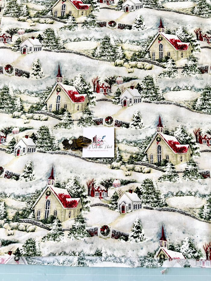 A close-up of a Susan Winget Country Church Christmas-themed fabric print featuring snow-covered hills, small houses, churches, trees, and wreaths in white, red, green, and beige tones. A small white card with a logo is placed over the center of the fabric.