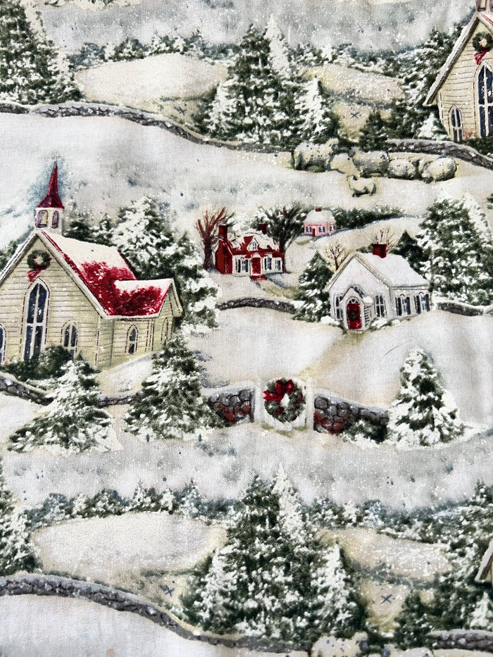 A close-up of a Susan Winget Christmas-themed fabric print featuring snow-covered hills, evergreen trees, small houses, and a church with red roofs and wreaths.