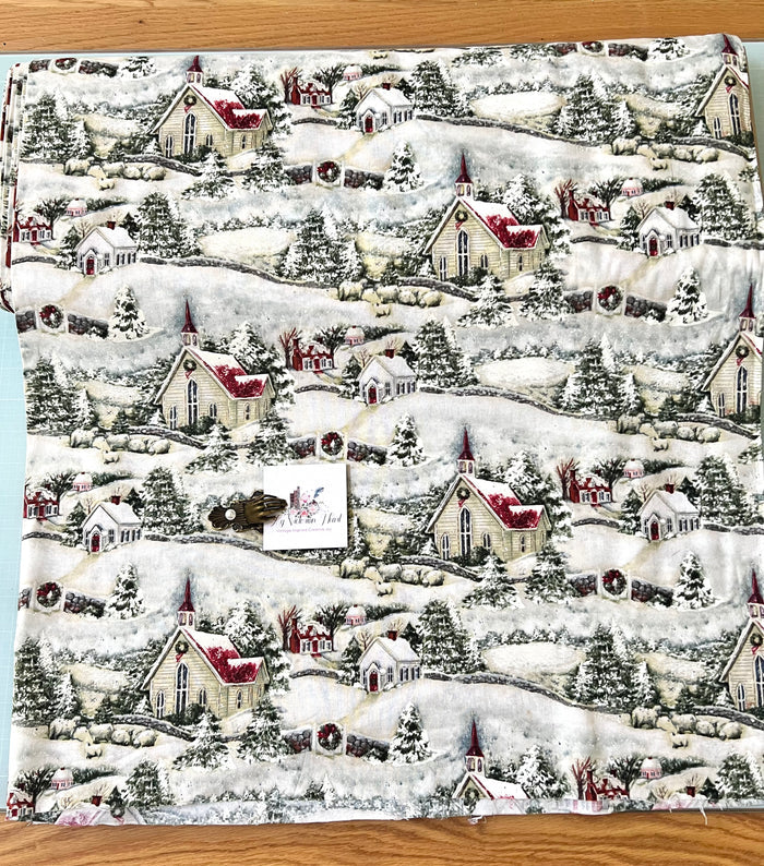 Susan Wi get Country Church Christmas fabric with a repeating Christmas village pattern featuring snow-covered houses, trees, and fences on a white background, laid flat on a wooden surface.