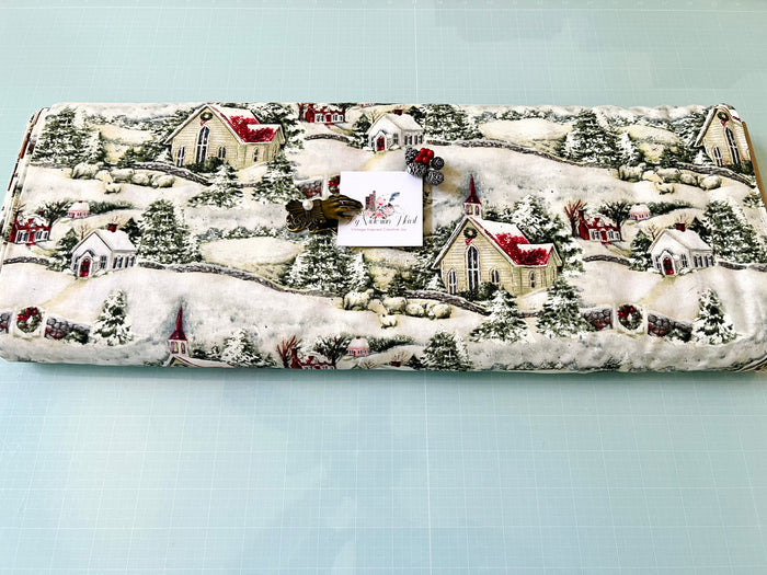 Bolt of Susan Wi get Country Church Christmas fabric featuring a winter village scene with snow-covered houses, trees, sheep, fences, garland, wreaths displayed on a light blue cutting mat.