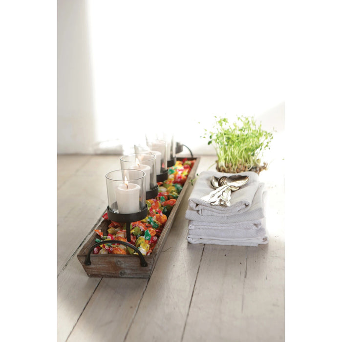 The CREATIVE COOP VOTIVE HOLDER features a rectangular wood and metal frame with handles, five white candles in glass votives on black stands, and decorative pebbles at the base—ideal for creating cozy fireplace ambience.